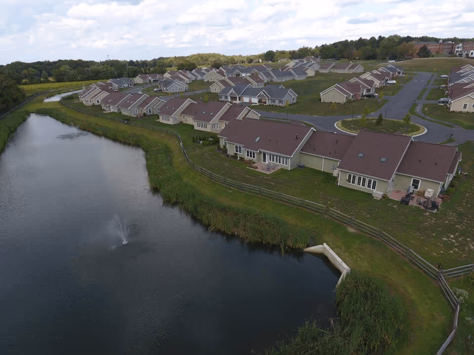 Aerial view of a residential community with multiple single-story houses arranged in rows near a pond with a small fountain. The area is surrounded by greenery and trees in the background.