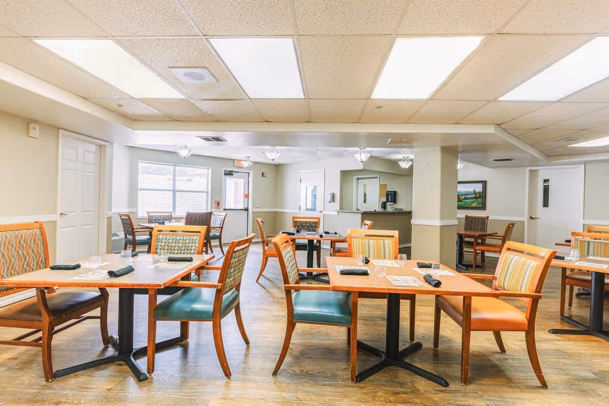 A bright dining room in a senior living facility with multiple wooden tables and chairs arranged neatly. Each table is set with placemats, napkins, and glassware. The room has large windows letting in natural light, light-colored walls, and a wood floor. There are doors and a counter area visible in the background.