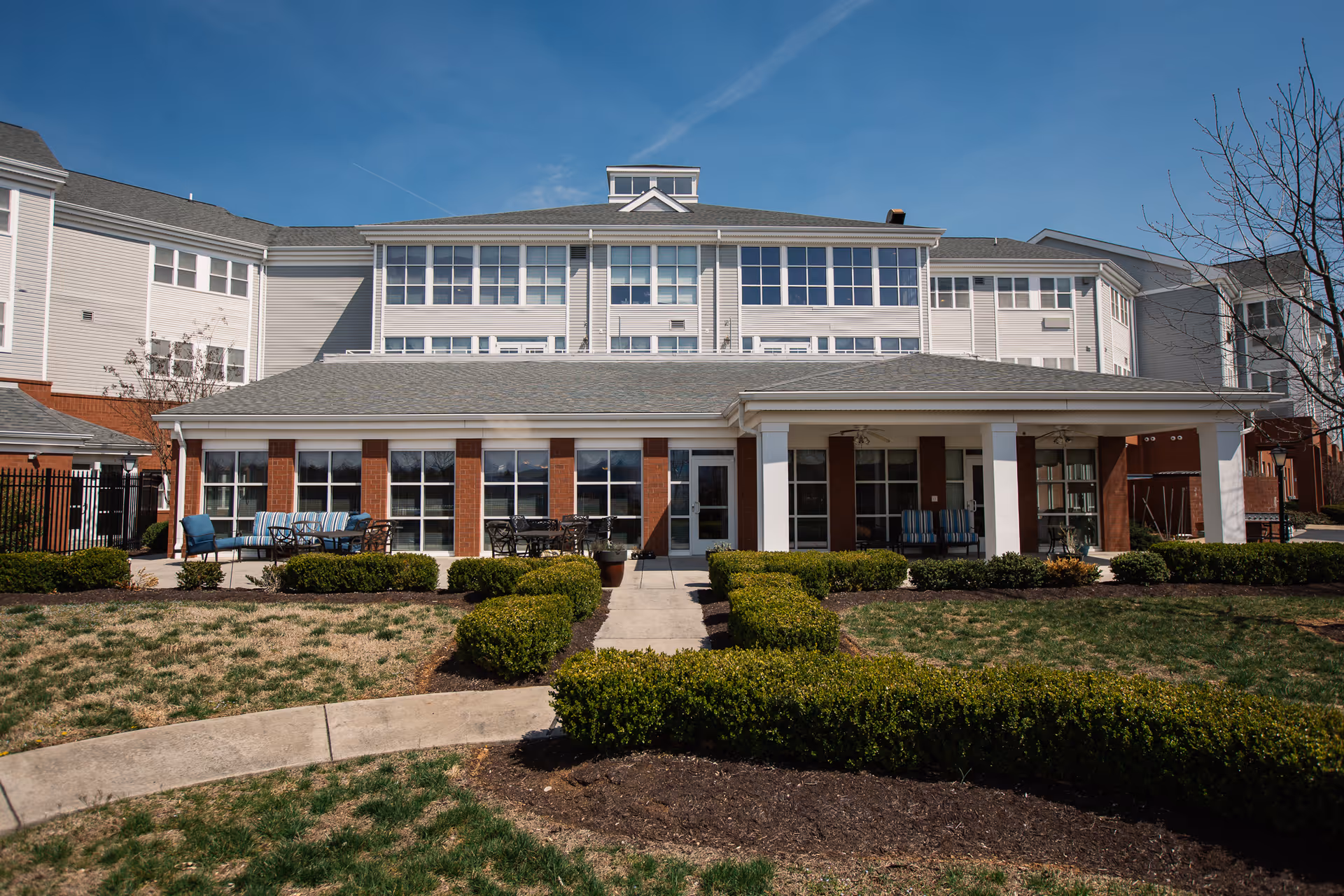 Exterior view of a multi-story senior living facility building with large windows and a covered patio area furnished with outdoor seating. The foreground features a landscaped garden with trimmed bushes and a concrete walkway leading to the entrance.