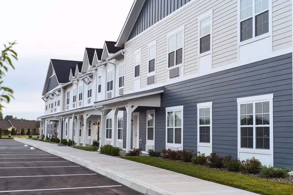 Front exterior of a two-story senior living building with multiple windows, a covered porch, landscaping, and an adjacent parking lot.