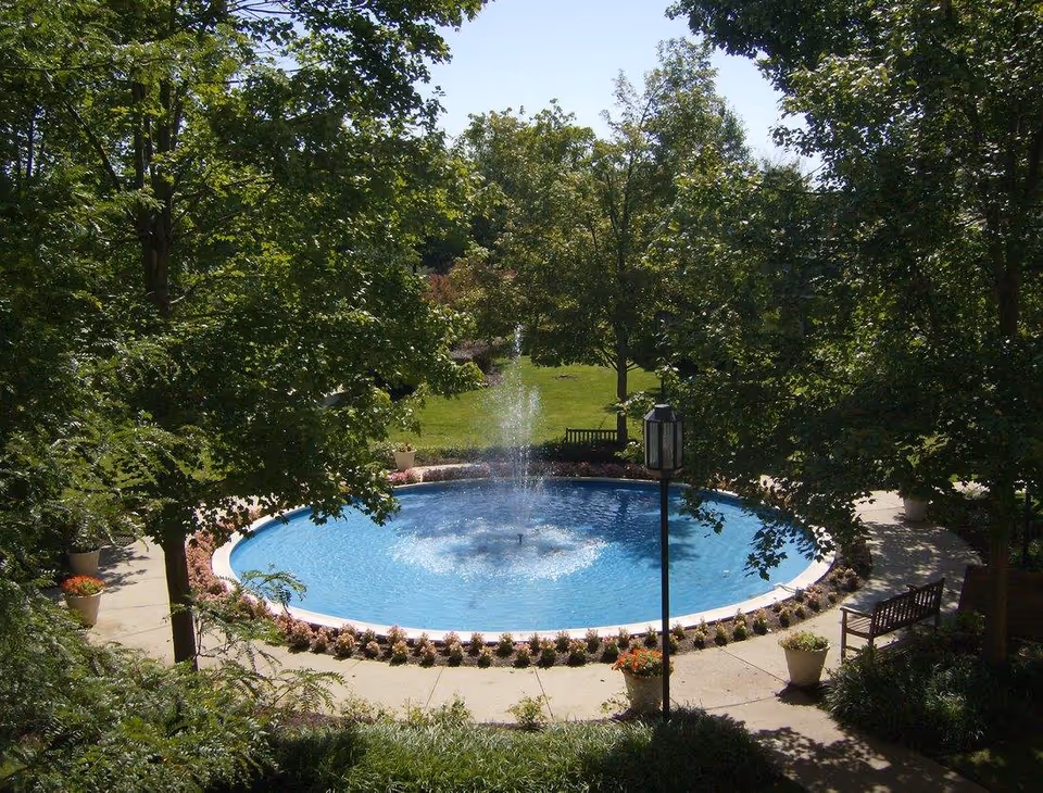 A circular water fountain surrounded by a paved walkway, benches, potted plants, and lush green trees in an outdoor garden area.