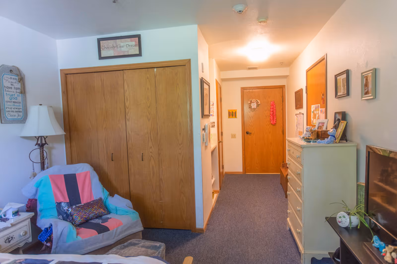 Interior view of a senior living facility room with a cozy armchair covered by a colorful blanket, a wooden closet with double doors, a white dresser with various items on top, framed pictures on the walls, and a hallway leading to a closed wooden door decorated with a wreath and a pink lei.