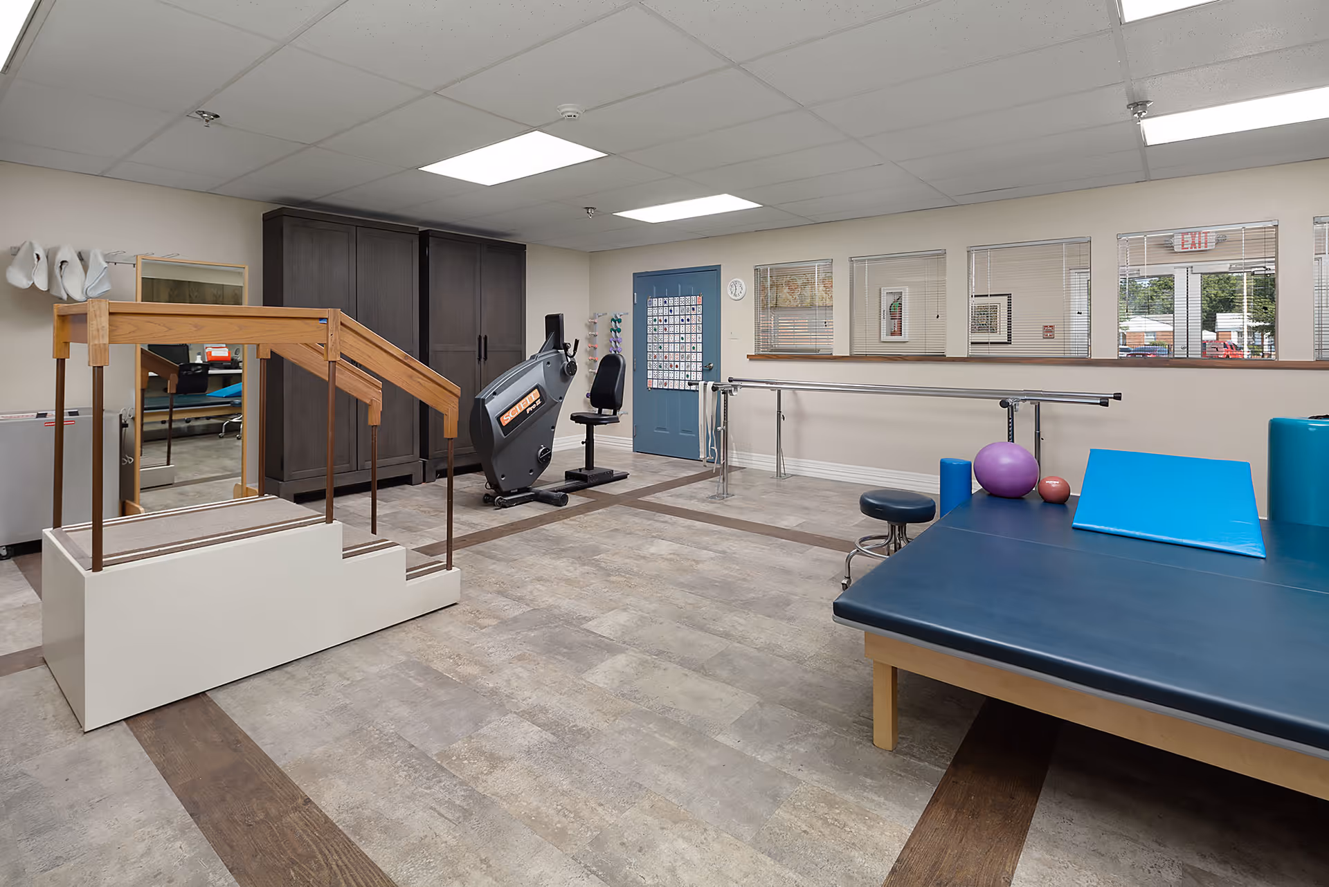 Rehabilitation therapy room with parallel bars, a recumbent exercise bike, a treatment table, therapy balls, and gait-training stairs.