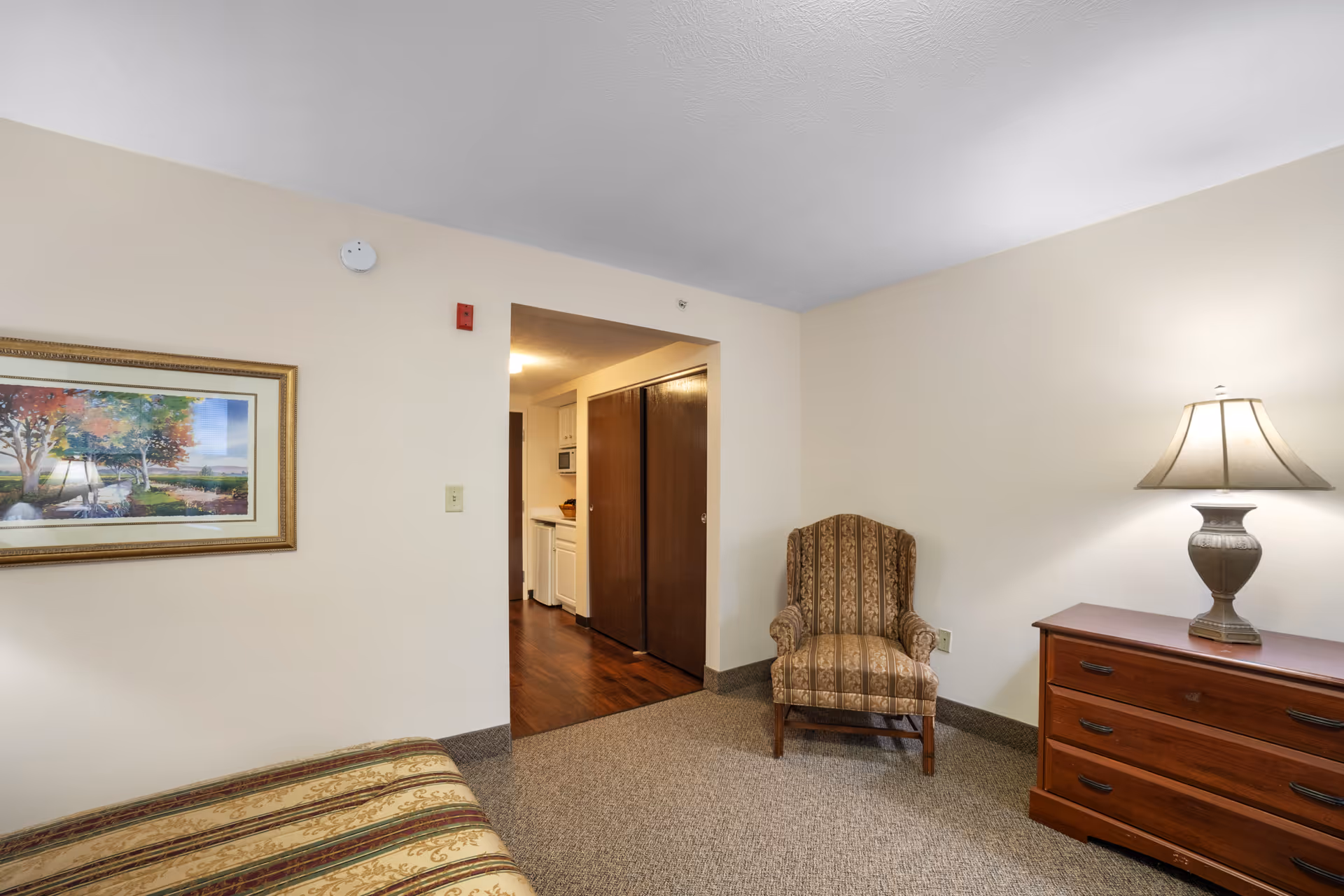 A cozy interior room with beige walls and carpeted floor. The room features a patterned armchair, a wooden dresser with a decorative lamp on top, and a framed landscape painting on the wall. An open doorway leads to a small kitchen area with white cabinets and wooden sliding doors.