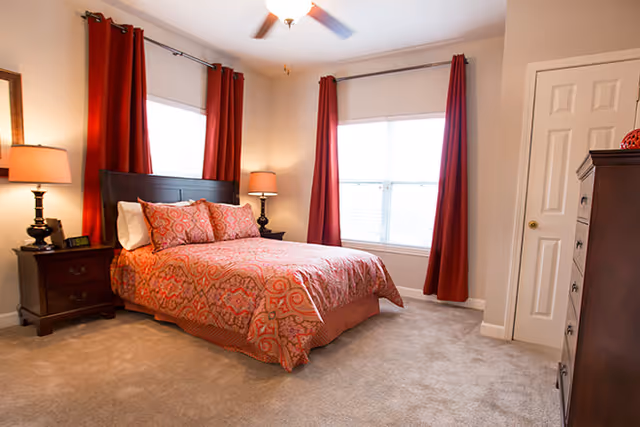 Bright bedroom with a queen bed dressed in patterned red bedding, matching red curtains, bedside tables with lamps, a dresser and a closet door.