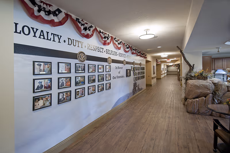 A long hallway in a senior living facility decorated with a patriotic theme honoring veterans. The left wall features framed photos of veterans under words like 'LOYALTY', 'DUTY', 'RESPECT', and 'SELFLESS SERVICE'. Red, white, and blue bunting decorates the top of the wall. The floor is wood-style, and the hallway extends into the distance with ceiling lights and some furniture and plants on the right side.