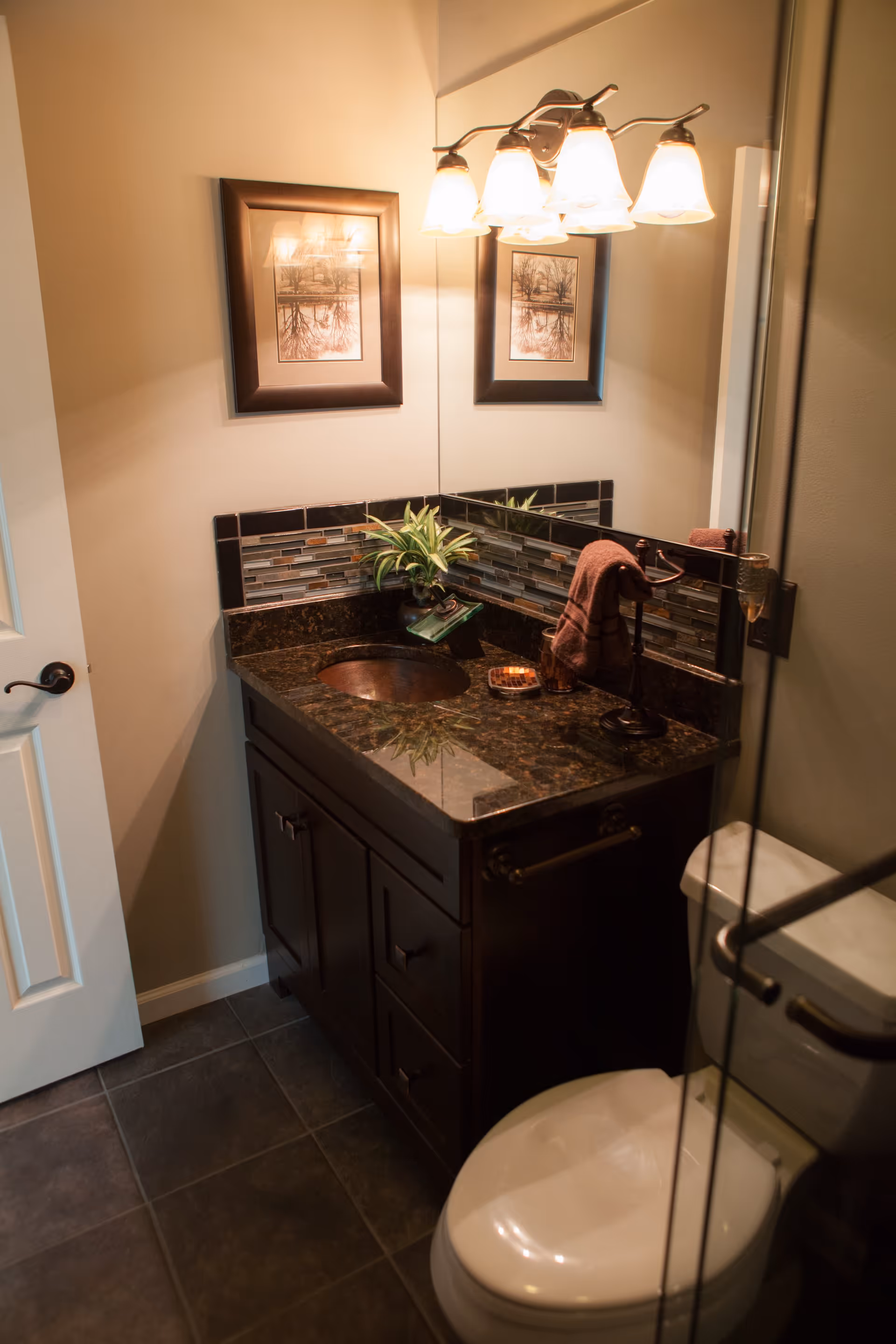 A small bathroom with a dark granite countertop vanity, a round sink, a toilet, and a large mirror above the vanity. The wall above the sink has a decorative tile backsplash and a framed picture. There is a light fixture with five lamps above the mirror, and a towel is hanging on a stand on the countertop.
