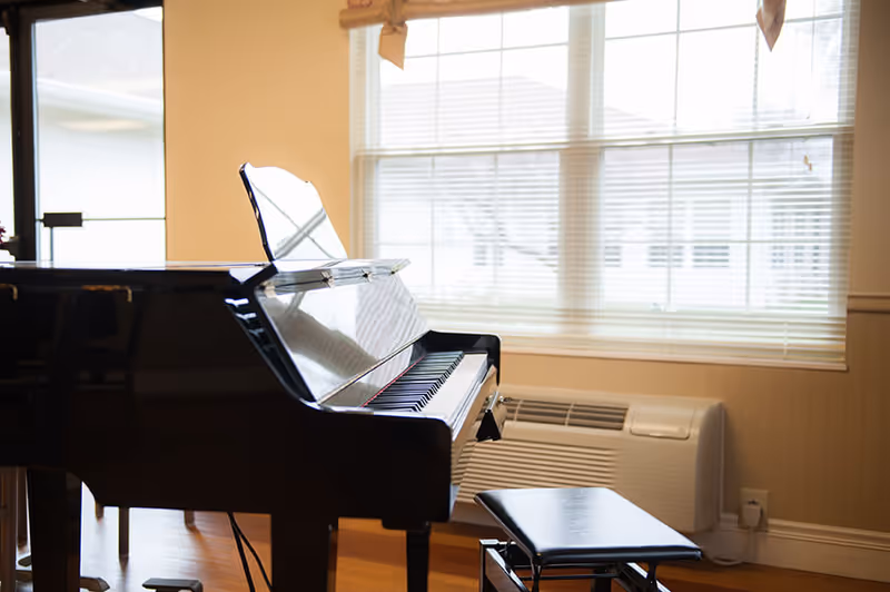 A black grand piano and bench sit in a bright room by a large window and a wall-mounted air conditioner.