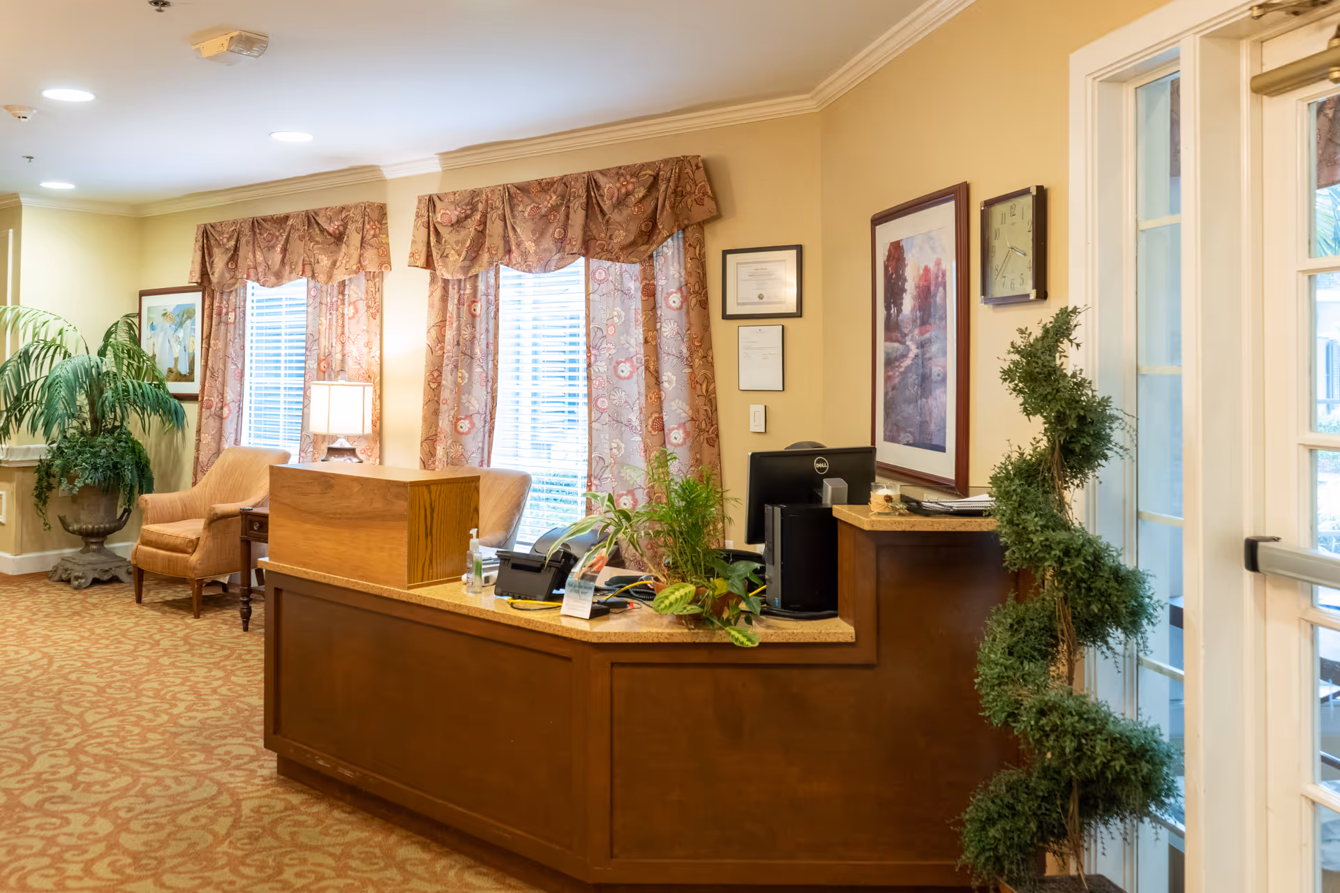 Reception area with a wooden front desk, computer, and plants. Behind the desk are two windows with floral curtains and a lamp on a side table between two upholstered chairs. The walls are decorated with framed artwork and a clock. A spiral-shaped topiary plant is near the glass door entrance.