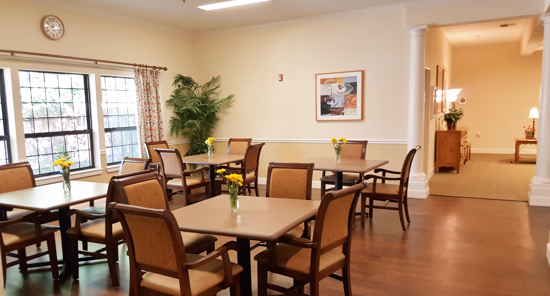 A bright dining room with several square tables, each surrounded by four wooden chairs with cushioned seats and backs. Small vases with yellow flowers are placed on each table. The room has large windows with floral curtains, a wall clock, a framed artwork, and a potted plant in the corner. The floor is wooden, and there is a hallway visible in the background with additional furniture and lamps.