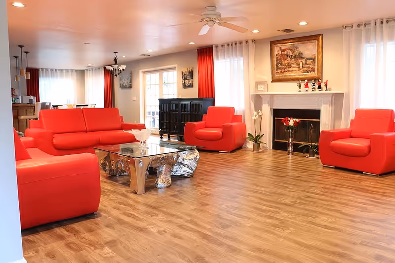 A bright living room with red leather sofas and armchairs arranged around a glass coffee table with a metallic base. The room features wood flooring, a white ceiling fan, a fireplace with decorative items on the mantel, and large windows with white and red curtains allowing natural light to fill the space.