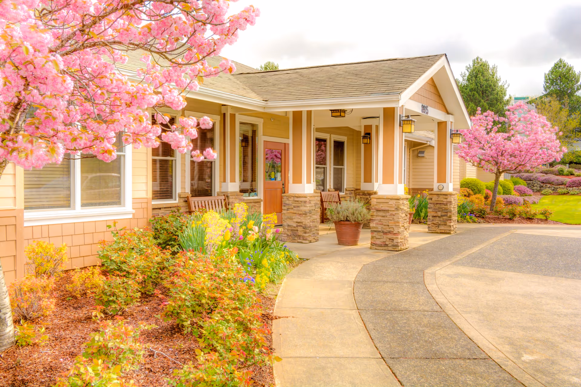 Entrance of a single-story building with a covered porch supported by stone pillars, surrounded by blooming pink cherry blossom trees and well-maintained garden beds with various flowers and shrubs.