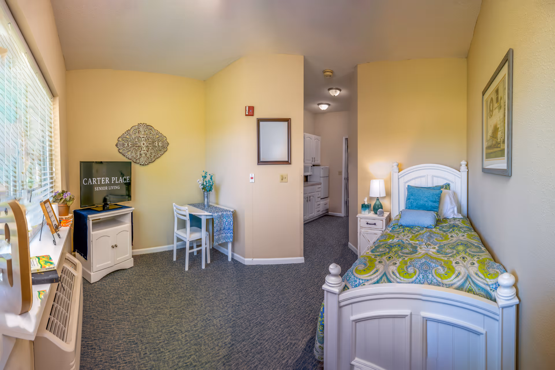 A small furnished senior-living bedroom with a single bed, nightstand and lamp, a TV reading 'Carter Place Senior Living', a small table and chairs, and a kitchenette visible in the background.