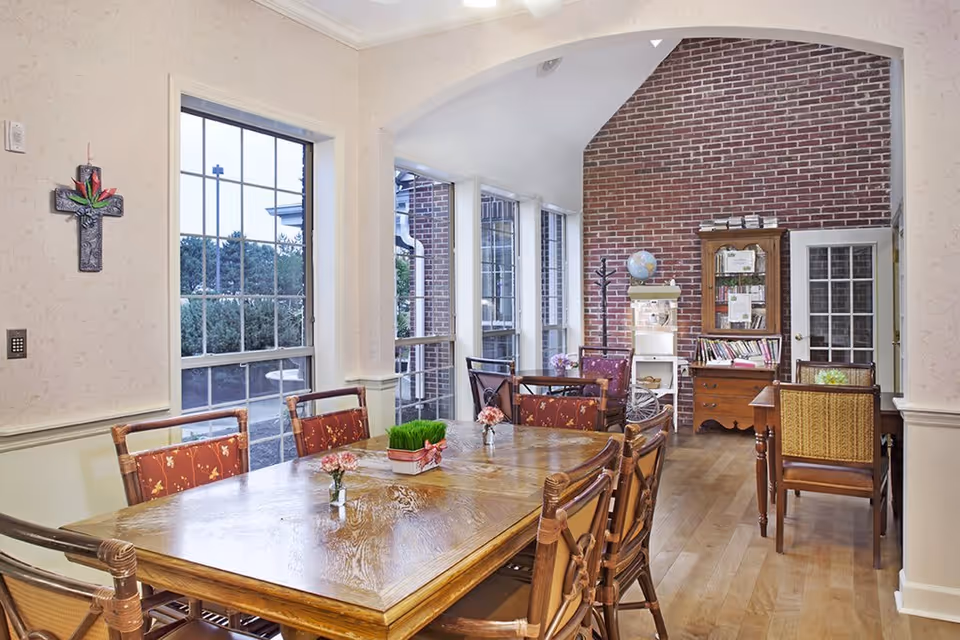 A bright dining room with a large wooden table surrounded by chairs with red patterned cushions. The room has large windows letting in natural light, a brick accent wall, a bookshelf filled with books, and a globe on a small white shelf. There is a decorative cross on the wall and wooden flooring throughout.