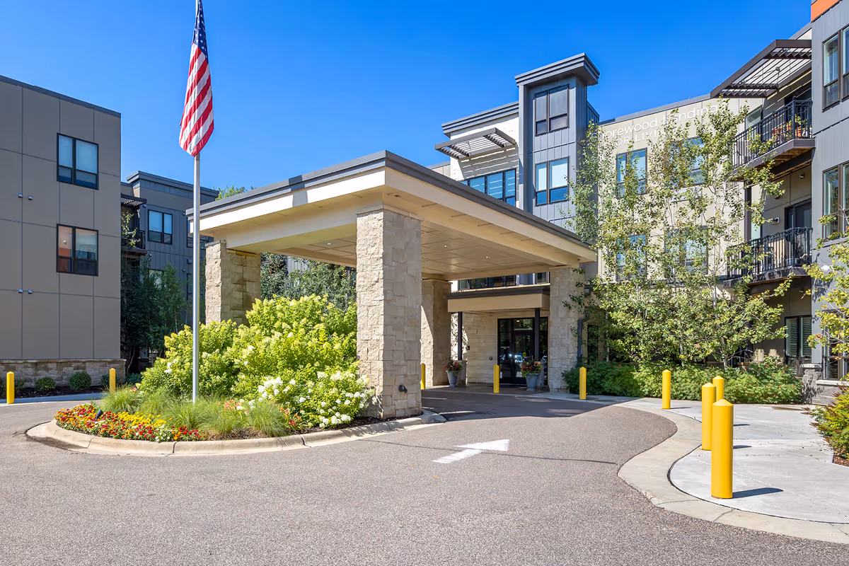Exterior view of Shorewood Landing senior living facility entrance with a covered drop-off area, landscaped greenery, an American flag on a flagpole, and a clear blue sky.