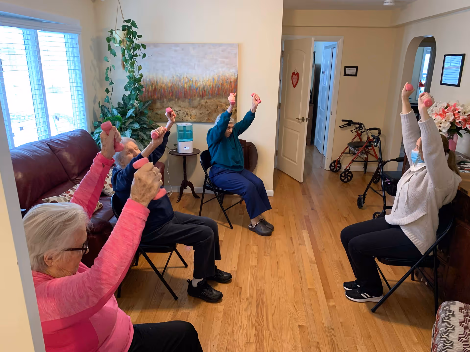 Four elderly individuals seated in a living room, participating in a seated exercise session using small pink hand weights. The room has wooden floors, a large window with blinds, a red leather couch, a green plant, a painting on the wall, and mobility walkers in the background.