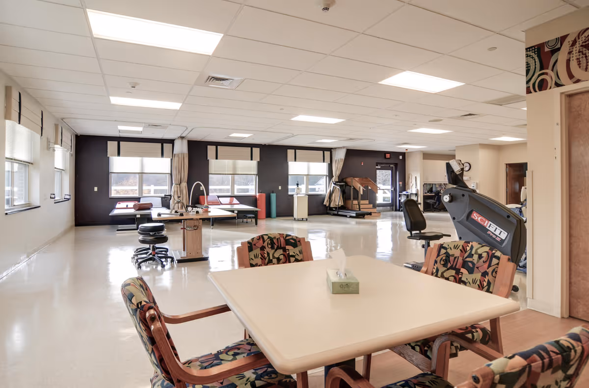 A spacious rehabilitation room with exercise equipment including a SciFit machine, therapy tables, and chairs around a square table with a tissue box on it. The room has large windows with blinds and curtains, a tiled floor, and a drop ceiling with fluorescent lights.