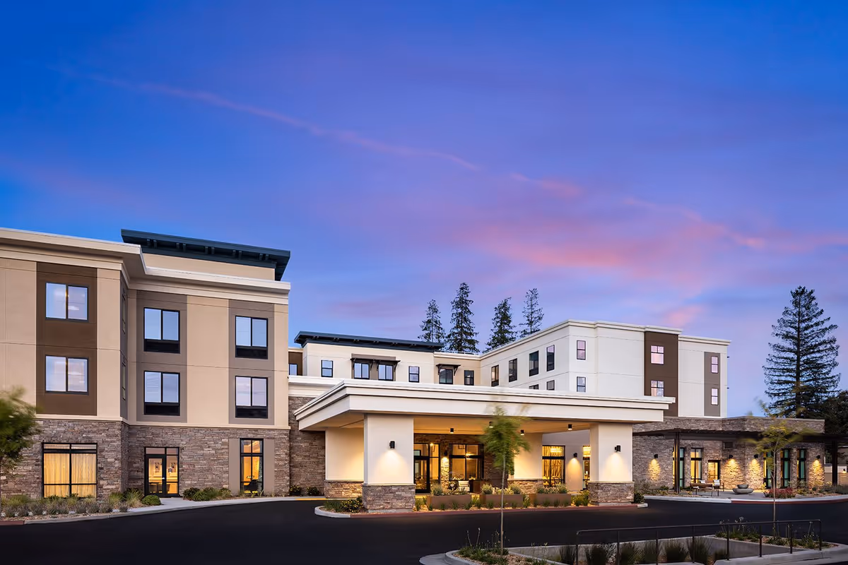 Exterior view of Belmont Village Senior Living Los Gatos building at dusk with a clear sky showing shades of blue and pink. The building has multiple floors with large windows, stone and beige walls, and a covered entrance with lights illuminating the facade. Trees and landscaped plants surround the entrance area.