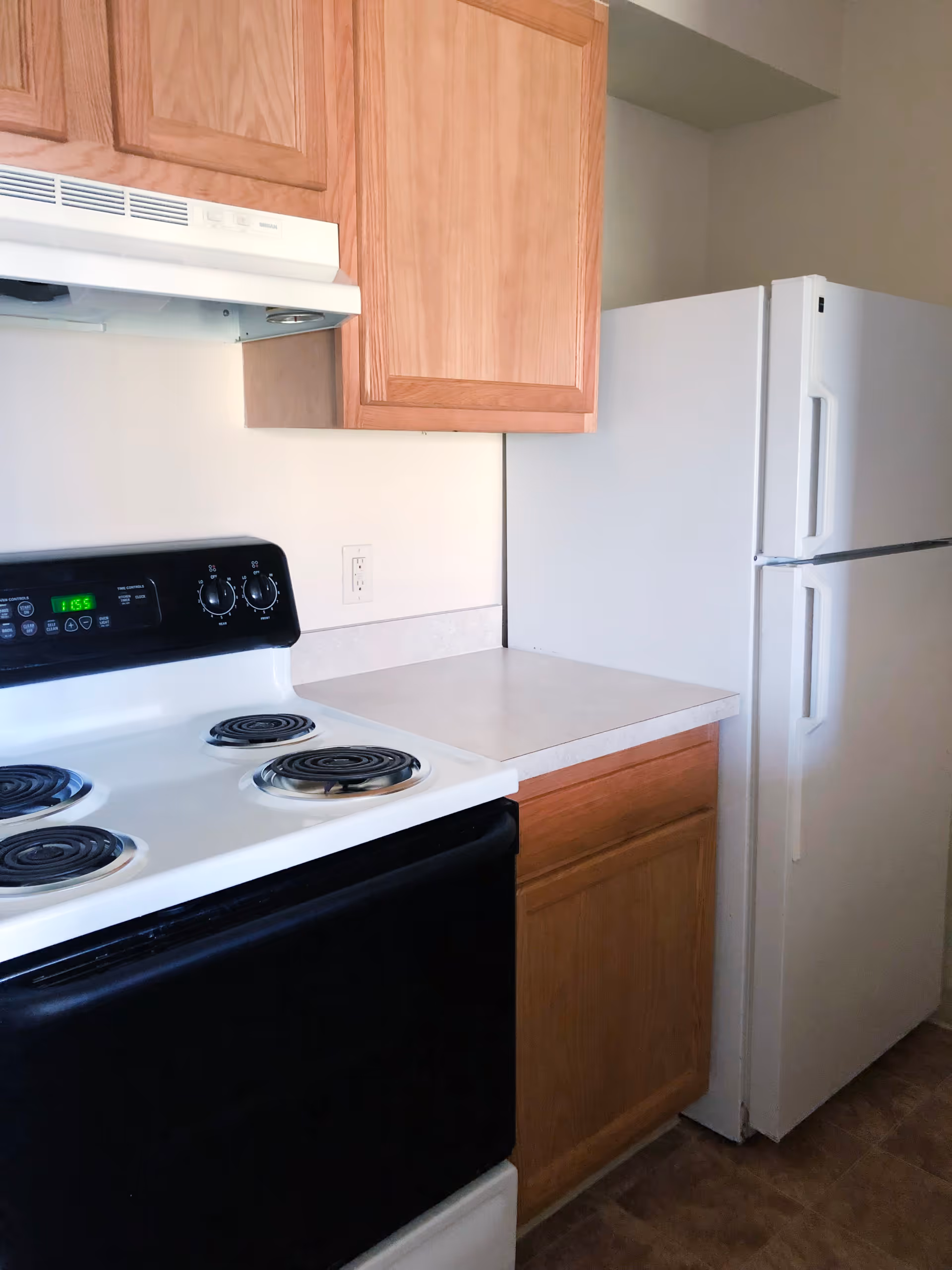 A kitchen corner featuring a white electric stove with black control panel and coil burners, a white refrigerator, light wood cabinets above and below a beige countertop, and a beige wall with an electrical outlet.