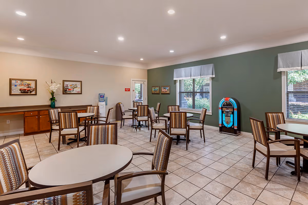 Bright communal dining area with round tables and chairs, tiled floor, framed art, windows, and a jukebox against a green accent wall.