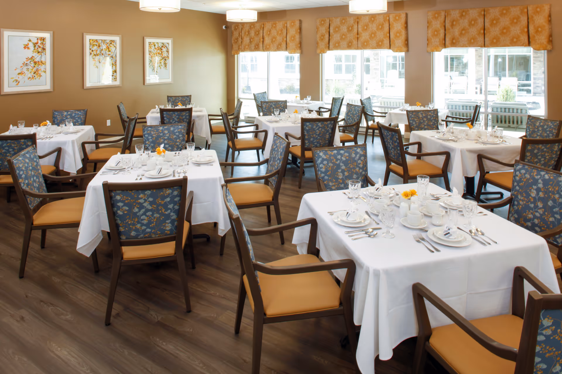 A bright and neatly arranged dining room with multiple square tables covered in white tablecloths. Each table is set with white plates, cups, glasses, and silverware. The chairs have wooden frames with floral-patterned backs and tan seats. Large windows with patterned valances allow natural light to fill the room, and framed floral artwork decorates the beige walls.