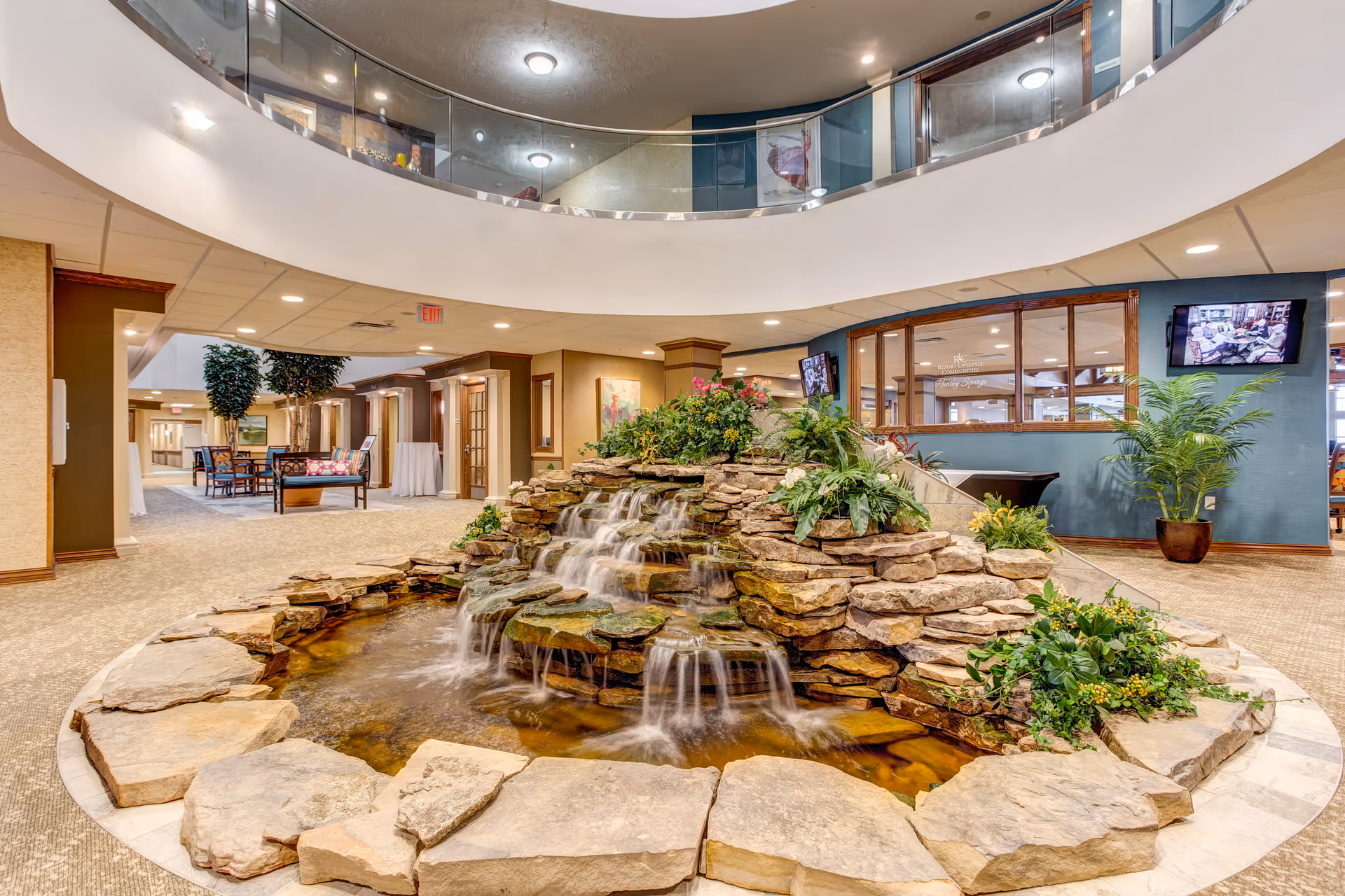 Indoor area of Pebble Springs Retirement Community featuring a decorative stone waterfall with flowing water surrounded by plants. The space has carpeted floors, seating areas with chairs and tables, and a second-floor balcony with glass railing overlooking the waterfall. Walls are adorned with artwork and there are two televisions mounted on the wall.