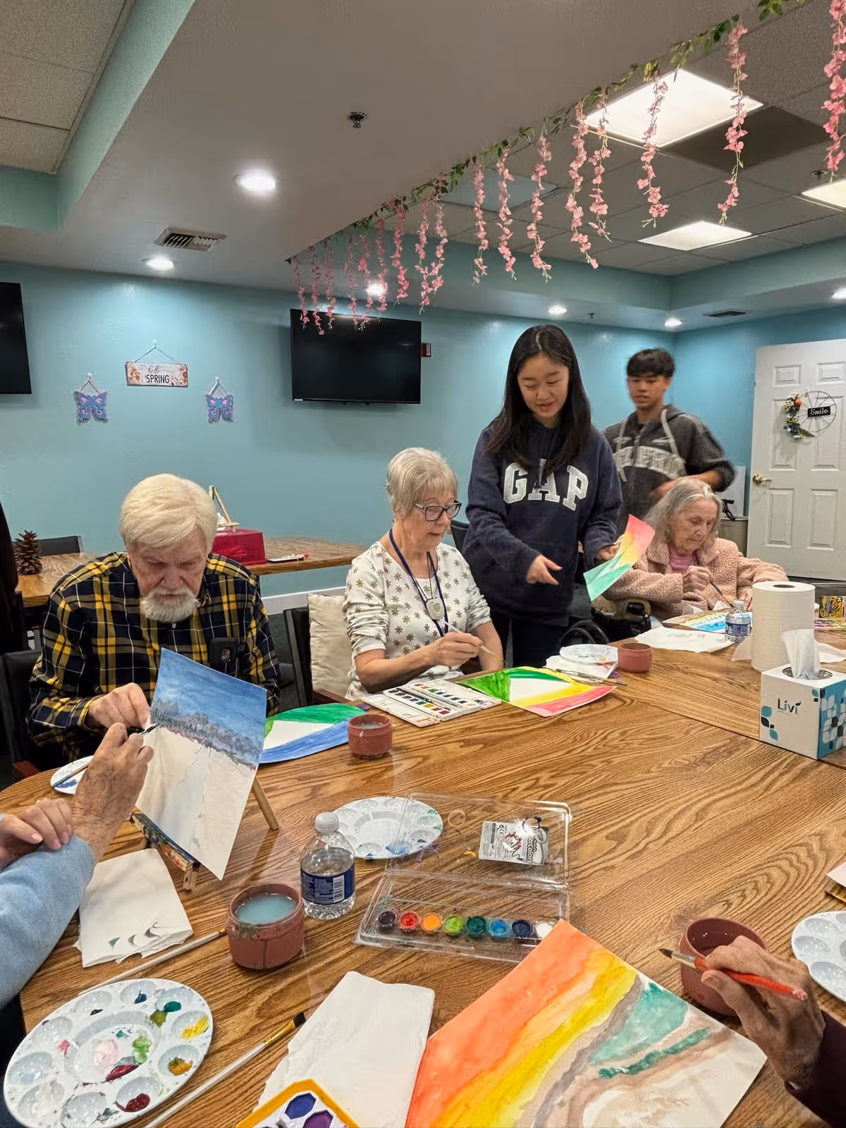 A group of elderly people sitting around a wooden table in a senior living community room, engaging in watercolor painting activities. Two younger individuals are standing nearby, assisting and interacting with the seniors. The room has light blue walls, a TV mounted on the wall, and pink hanging decorations from the ceiling.