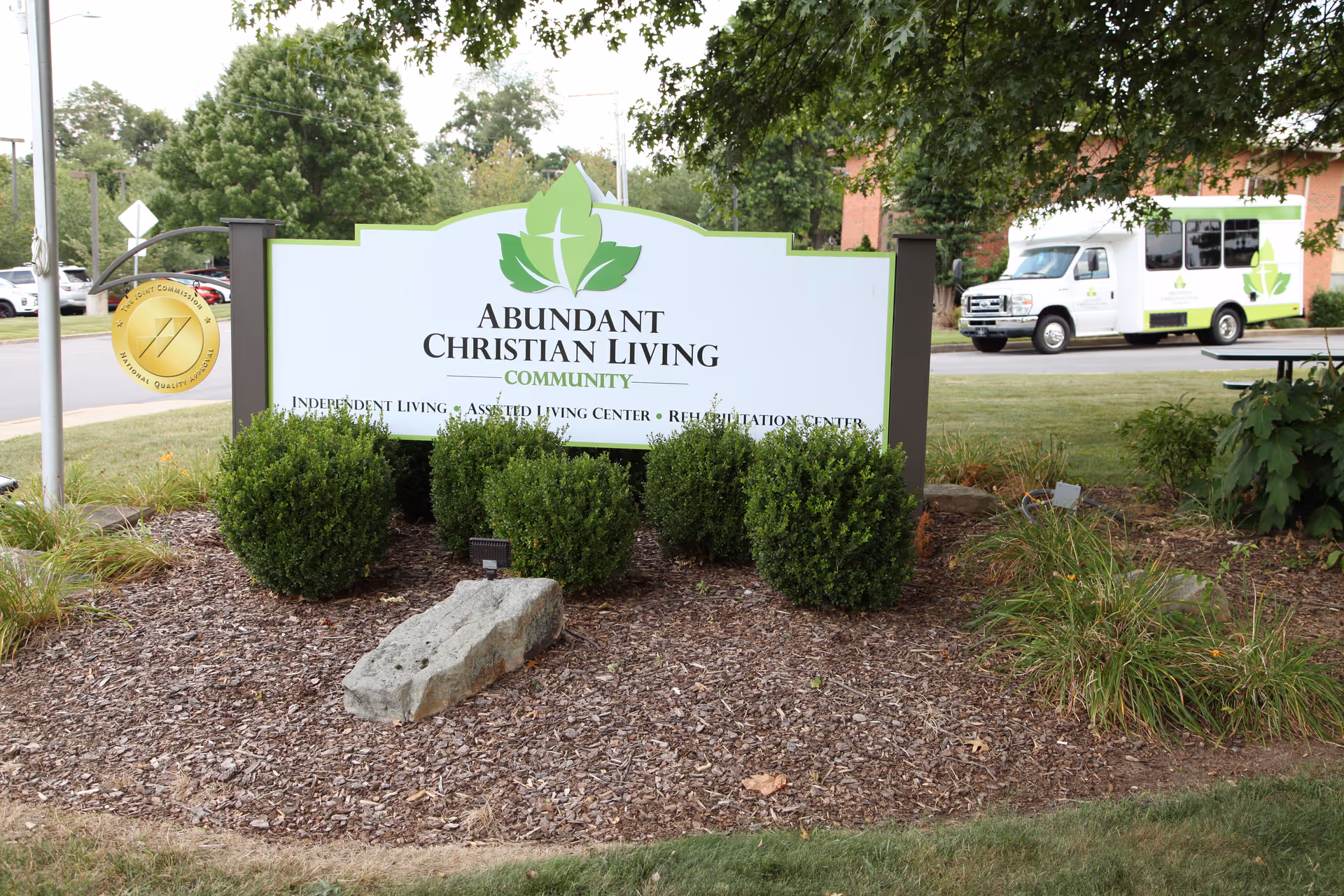 Outdoor view of a sign for Abundant Christian Living Community surrounded by bushes and mulch, with a white shuttle van and a brick building in the background.
