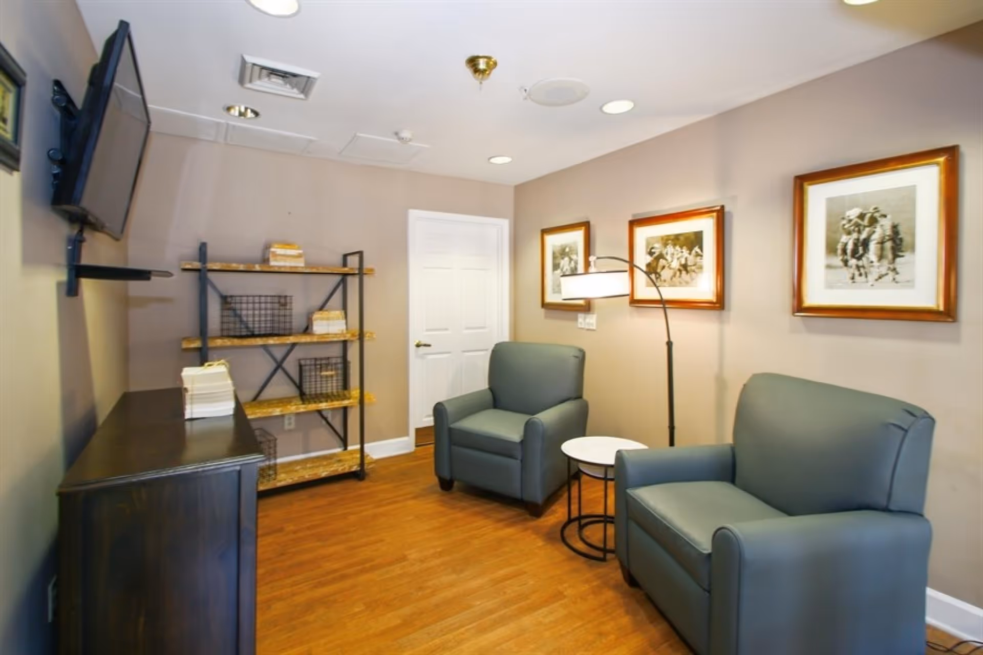 A cozy sitting area in a senior living community featuring two green armchairs separated by a small round white table with a black metal base. Behind the chairs, there is a floor lamp with a curved arm and a white lampshade. The walls are beige and decorated with three framed black and white pictures. On the left side, there is a wooden shelving unit with metal supports and some books and baskets on the shelves. A flat-screen TV is mounted on the wall above a dark wooden cabinet. The floor is wooden, and there is a white door in the background.