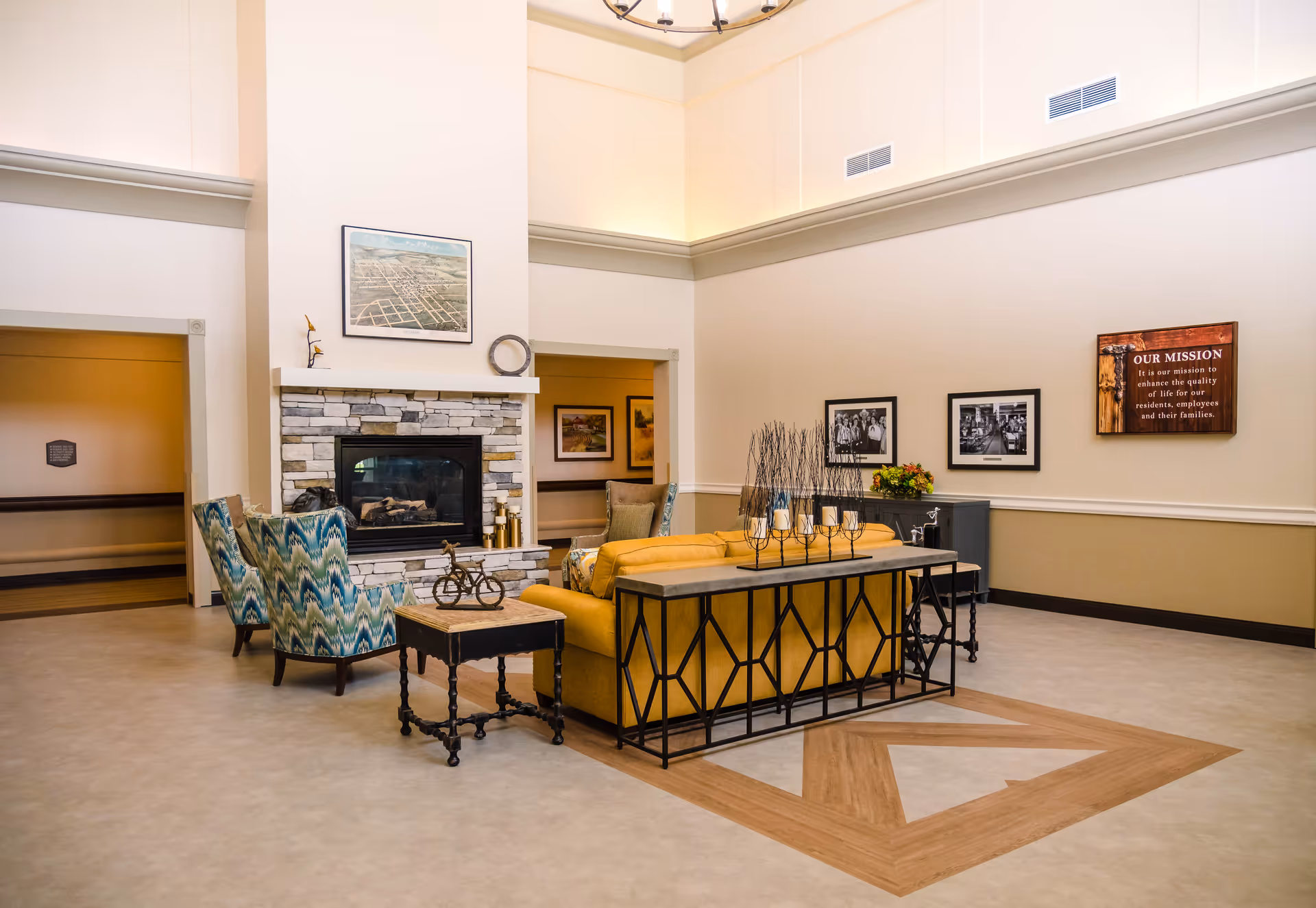 Spacious senior living room lounge with a yellow sofa, patterned armchairs, a stone fireplace, and framed wall decor.