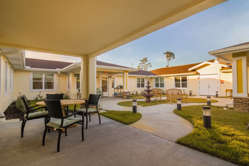 Outdoor courtyard area of a senior living facility with a covered patio featuring a round wooden table and four chairs with cushions. The courtyard has a paved walkway, green grass, a three-tiered fountain, and wooden benches. The building surrounding the courtyard is single-story with light yellow walls and white trim under a clear sky.