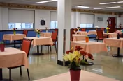A spacious dining room with multiple tables covered in peach-colored tablecloths, each table decorated with a small flower arrangement. The room has large windows with blinds, red chairs, and a clean, polished floor. There are white pillars and fluorescent ceiling lights.