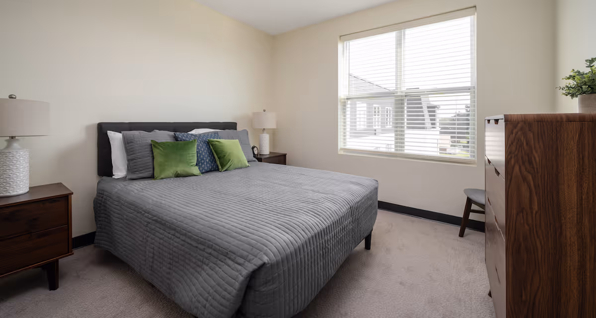Well-lit bedroom with a queen bed dressed in gray bedding and green accent pillows, bedside tables with lamps, a window with blinds, and a wooden dresser.