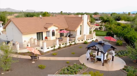 A single-story assisted living facility building with a tiled roof and a surrounding outdoor area featuring pathways, benches, umbrellas, and a gazebo with seating. The landscape includes small plants and trees, with mountains visible in the background under a clear sky.