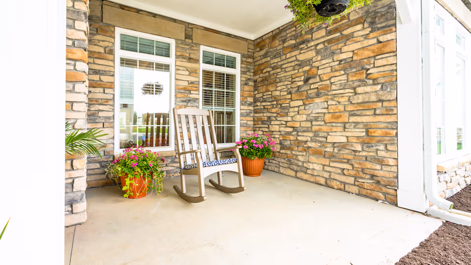 A covered outdoor patio area with stone walls, two windows, and a white rocking chair with a patterned cushion. There are two potted plants with pink flowers placed on the floor near the windows and a hanging plant above.