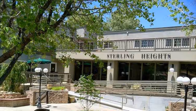 Exterior view of Sterling Heights Assisted Living facility showing a two-story building with a balcony, surrounded by trees and outdoor lamps. There is a ramp leading to the entrance and a patio area with seating and umbrellas.
