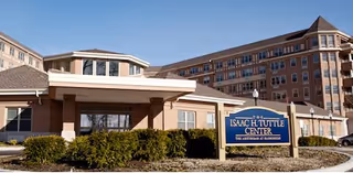 Exterior view of The Isaac H. Tuttle Center, a multi-story building with beige and brown tones, featuring a sign in front surrounded by bushes and a clear blue sky.