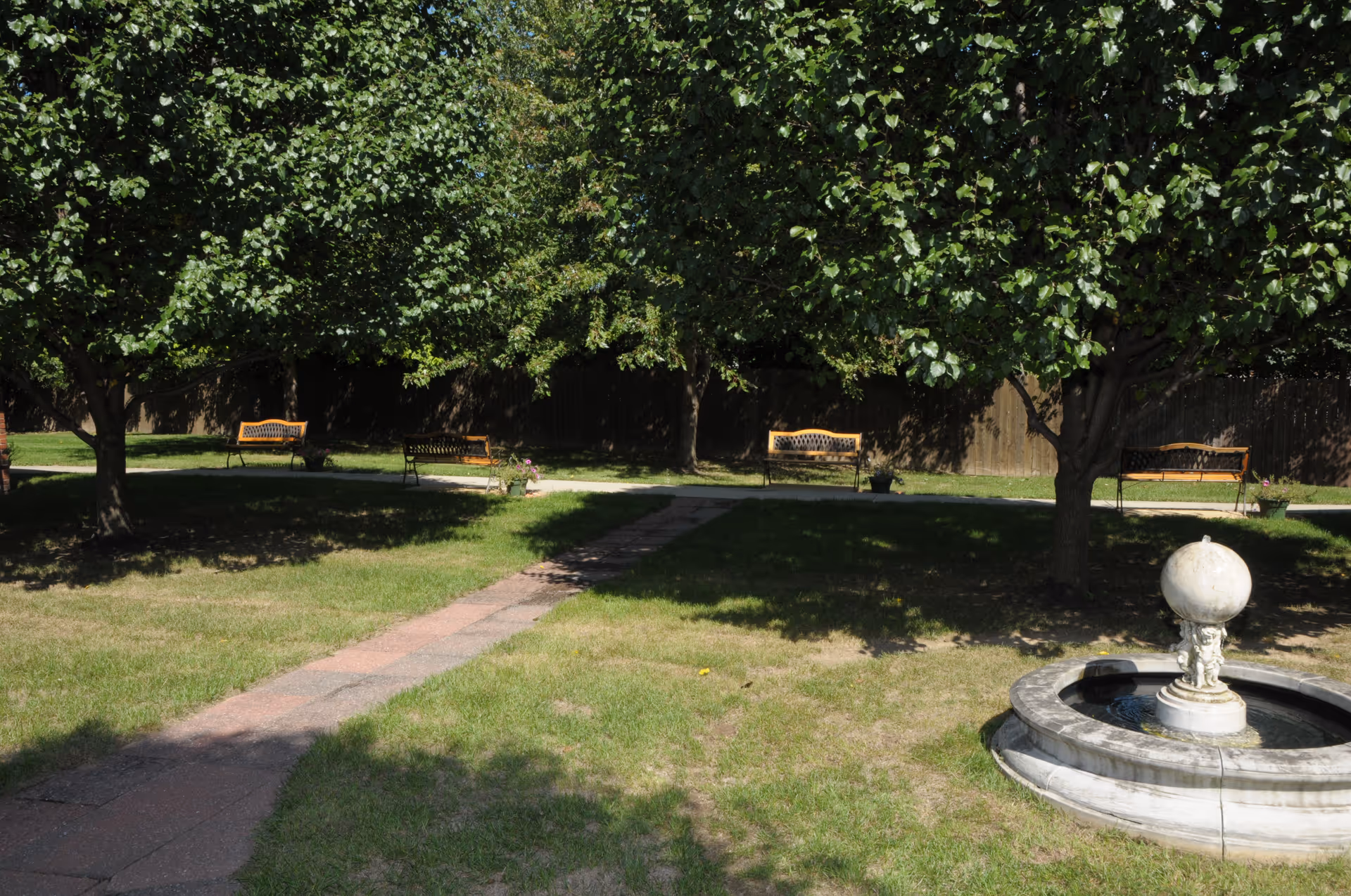 A peaceful outdoor garden area with a stone fountain in the foreground, several trees providing shade, and multiple wooden benches along a paved pathway.