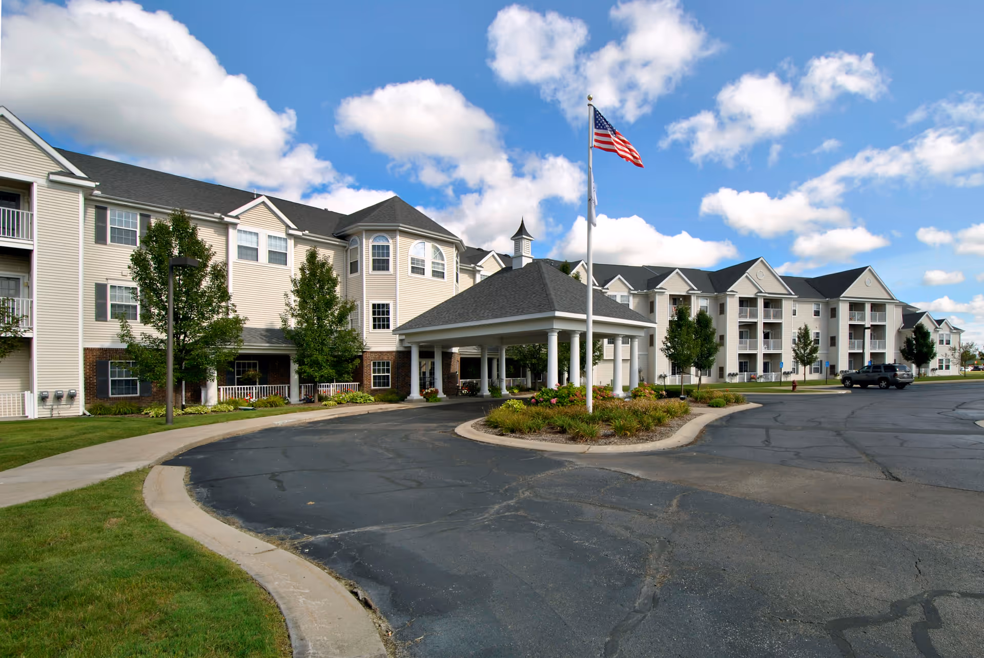 Exterior view of a large, multi-story retirement community building with beige siding and dark roofs under a partly cloudy blue sky. There is a covered entrance with white columns and an American flag on a flagpole in front. The driveway curves around landscaped areas with grass and trees.