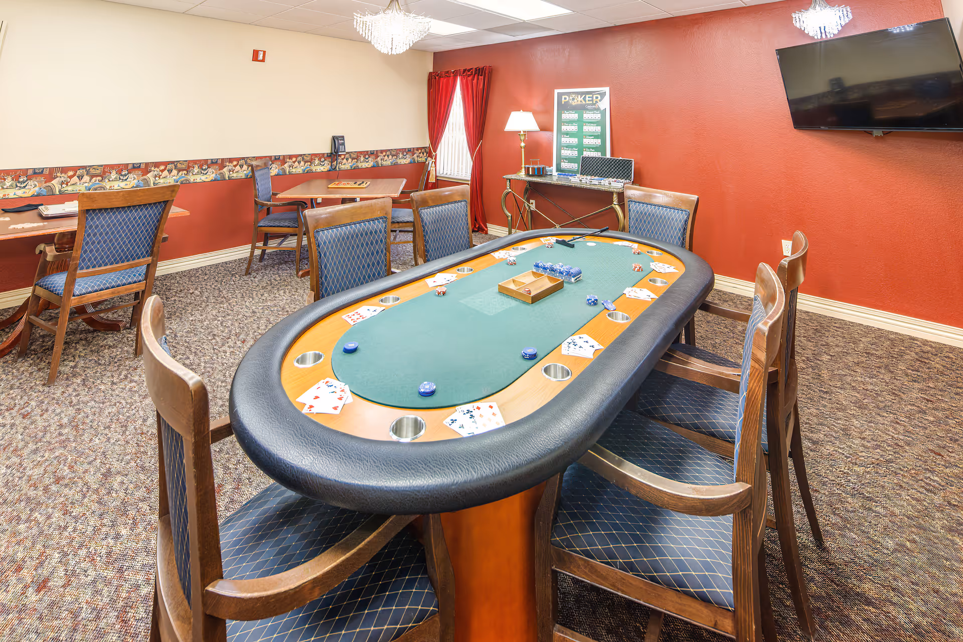 Poker table surrounded by chairs in a carpeted game room with red walls, a TV, and additional tables.