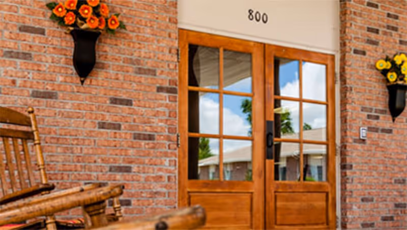Entrance to a building with double wooden doors featuring glass panes, set in a brick wall. The number 800 is displayed above the doors. Two black wall-mounted flower pots with orange and yellow flowers flank the doors. A wooden rocking chair is partially visible on the left side of the image.