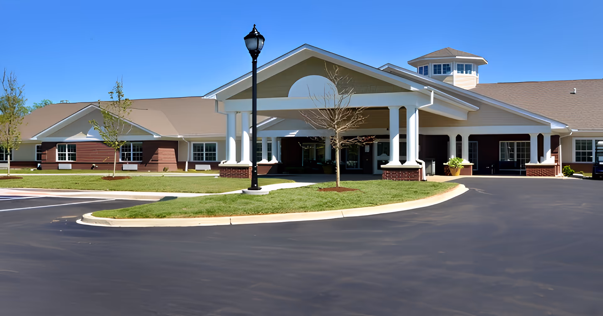 Front exterior view of The Lakes of Sylvania facility showing a large covered entrance with white columns, a paved driveway, manicured lawn, young trees, and a clear blue sky.