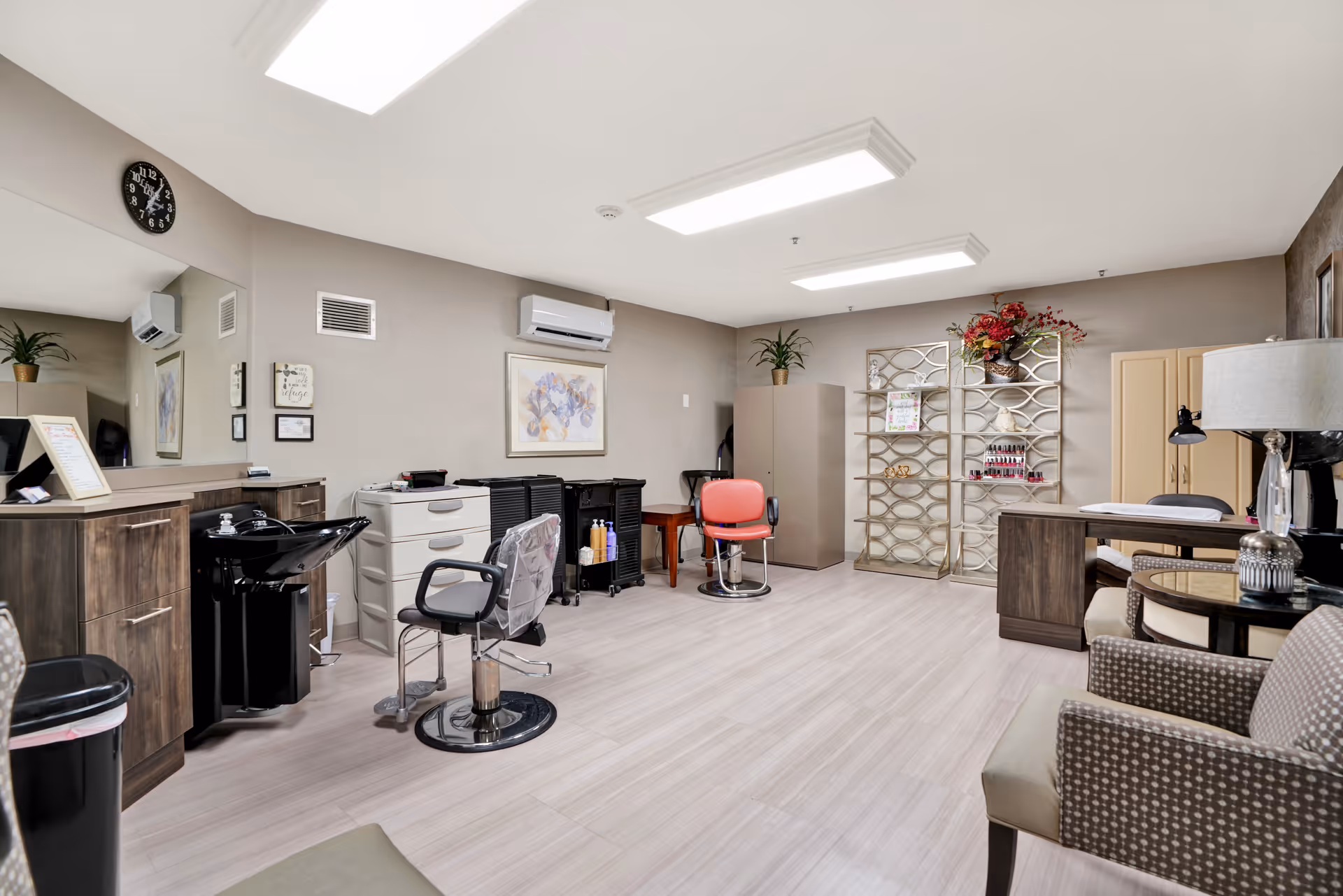 Interior view of a salon area in a senior living facility with two salon chairs, a hair washing station, cabinets, a clock on the wall, and decorative plants and artwork. There is also a nail polish display rack and a small seating area with armchairs and a side table with a lamp.