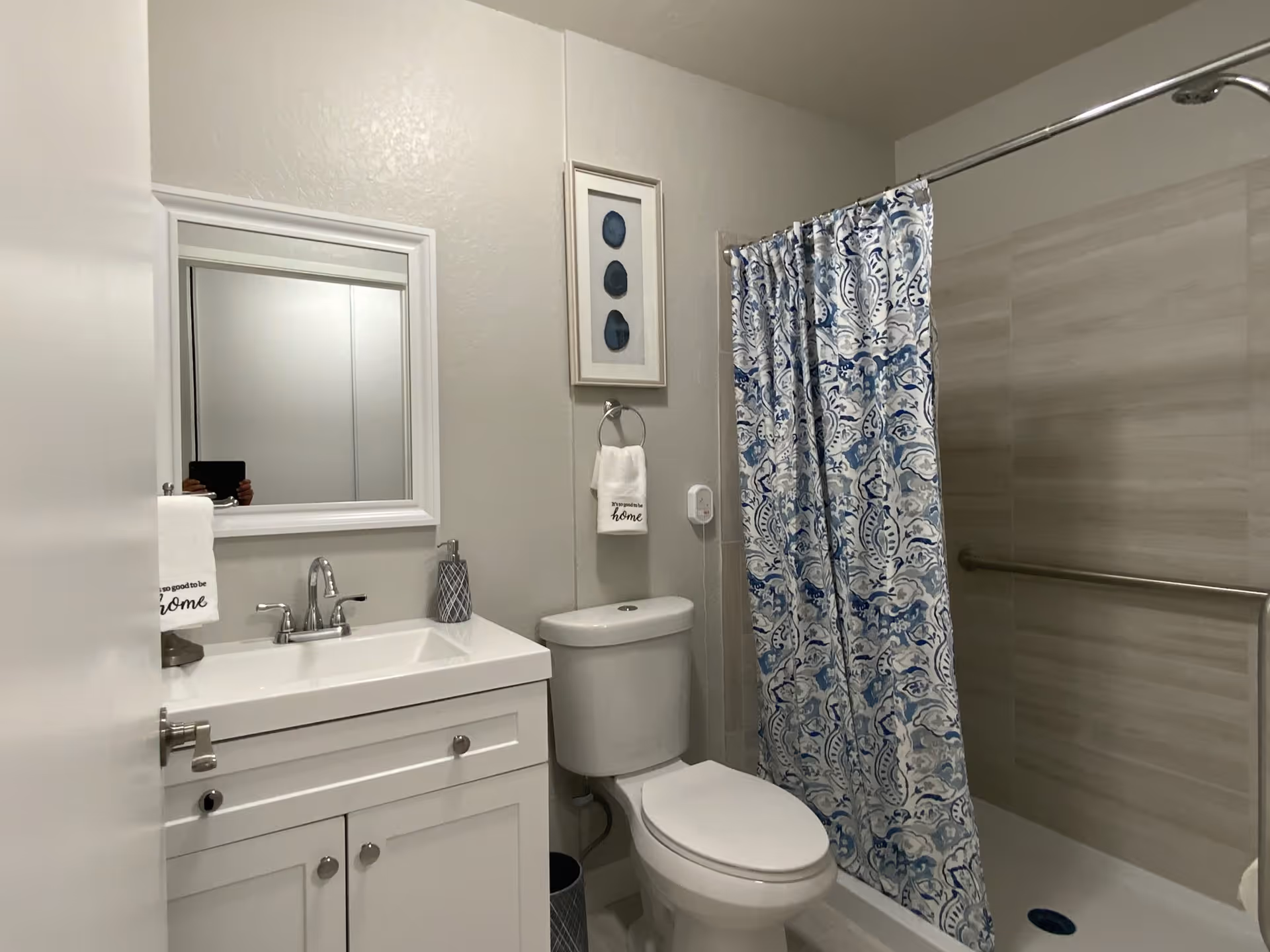 A clean and modern bathroom with a white vanity and sink, a mirror above the sink, a toilet, and a shower with a blue and white patterned curtain. There is a towel hanging on a ring with the text 'It's so good to be home' and a framed artwork on the wall.