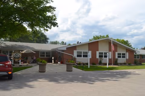 Exterior view of a single-story brick building with a sloped roof, an American flag on a pole, and a paved driveway with two large planters. There is a red vehicle parked on the left side and some outdoor seating with umbrellas near the entrance. Trees and a partly cloudy sky are visible in the background.