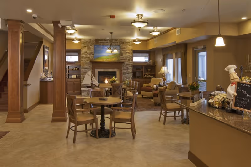A cozy senior living facility common area with wooden tables and chairs, a stone fireplace with a painting above it, comfortable armchairs, warm lighting, and a counter displaying a chef figurine and a chalkboard menu listing creamy potato soup.