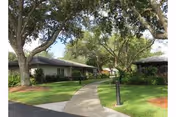 A paved walkway winding through a green lawn with large trees and shrubs, leading to single-story buildings with covered porches under a clear sky.