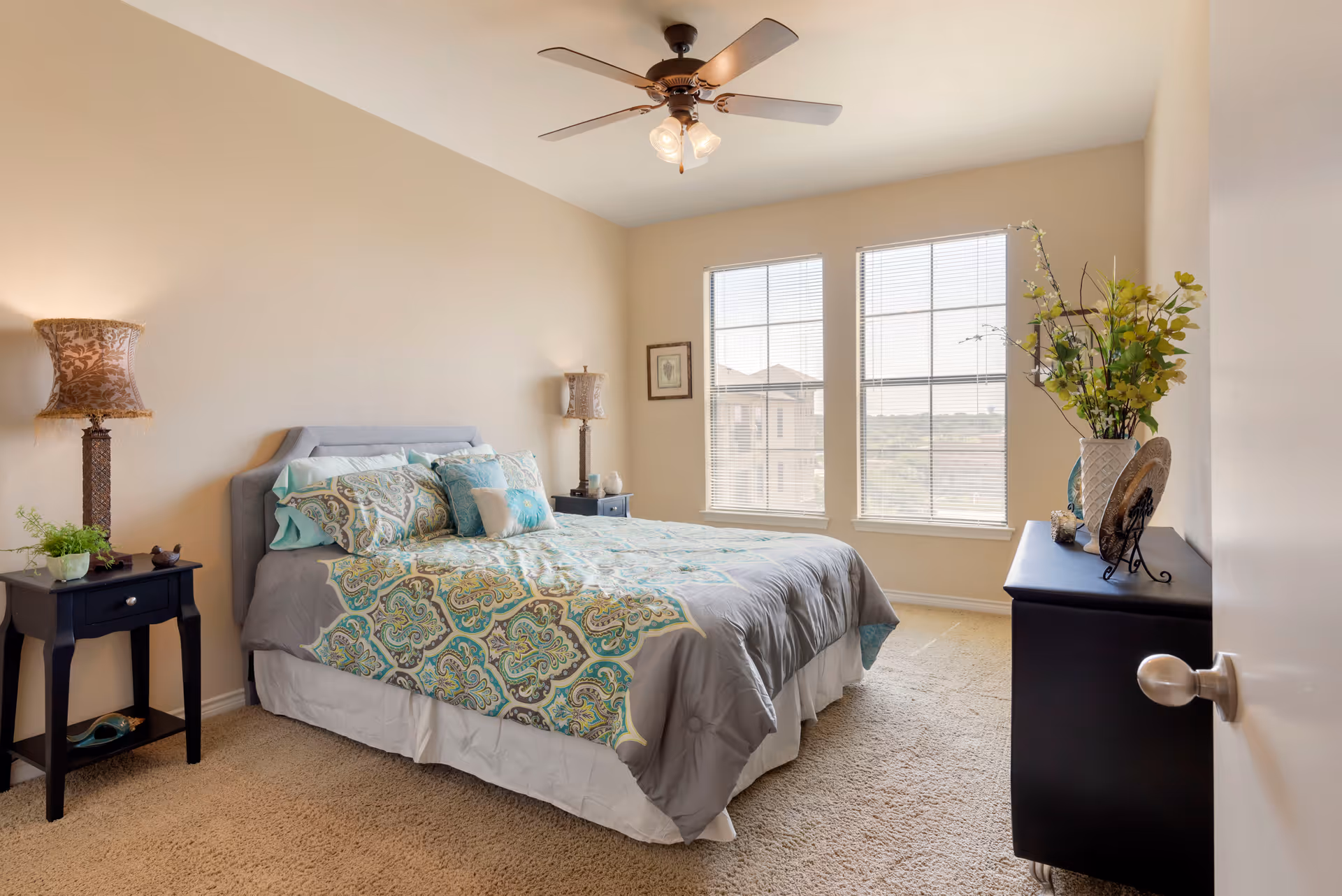 A bright bedroom with a large bed covered in patterned bedding in shades of blue, green, and gray. There are two bedside tables with decorative lamps and a small plant on one side. The room has beige walls, carpeted floor, a ceiling fan with lights, and two large windows letting in natural light. A black dresser with a vase of flowers and decorative items is positioned against one wall.