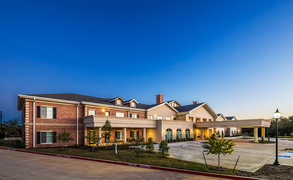 Exterior view of HarborChase of Southlake senior living facility at dusk, showing a large two-story building with brick and beige siding, a covered entrance, landscaped greenery, and a clear sky.