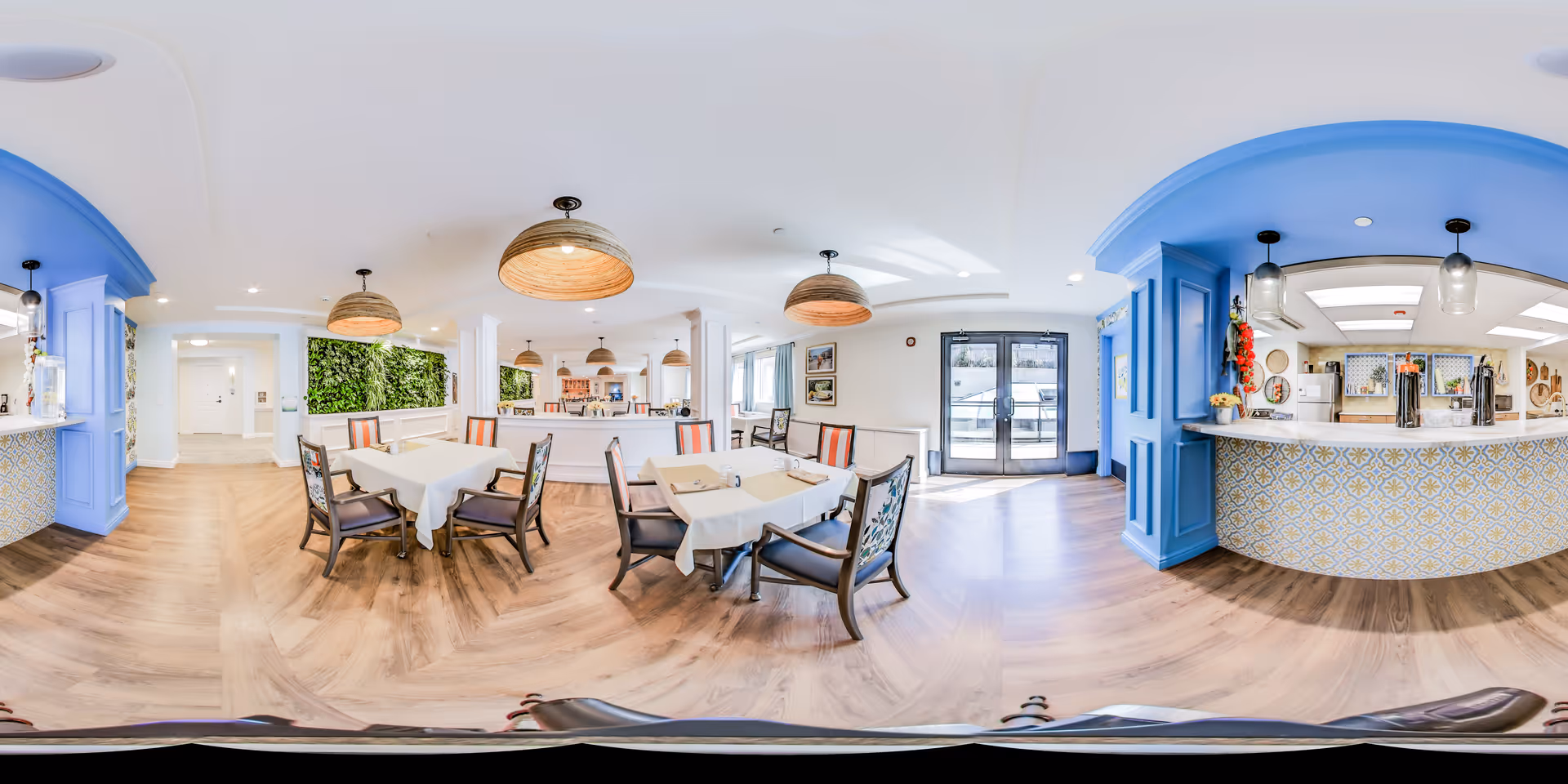 Bright and spacious dining area with several tables covered in white tablecloths and surrounded by chairs. The room features wooden flooring, blue archways, hanging pendant lights with woven shades, and a counter area with patterned tiles. There is a green plant wall on one side and large glass doors letting in natural light.